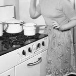 black and white photo of retro housewife standing at vintage stove
