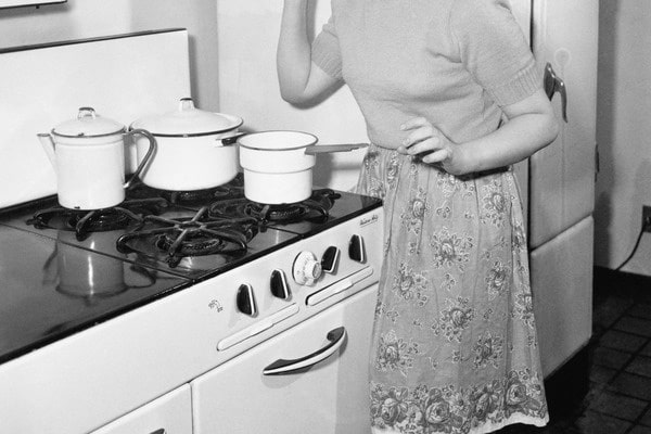 black and white photo of retro housewife standing at vintage stove