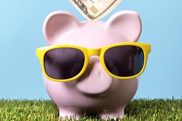 Hand putting a ten dollar bill into a pink piggy bank with sunglasses grass and blue sky