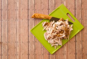 Shredded rotisserie chicken on a green plastic cutting board and carving knife against wood plank background