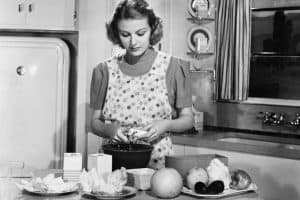 black and white photo of 1940s housewife wearing apron standing at kitchen counter prepping food